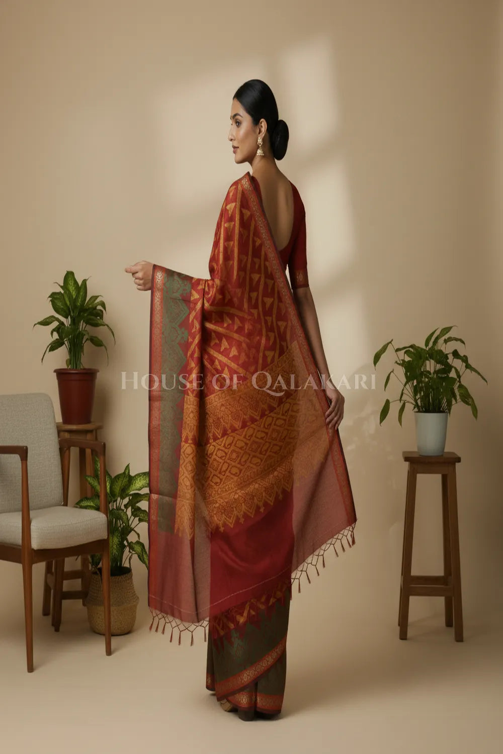 Woman in a patterned saree standing in a room with plants and furniture.